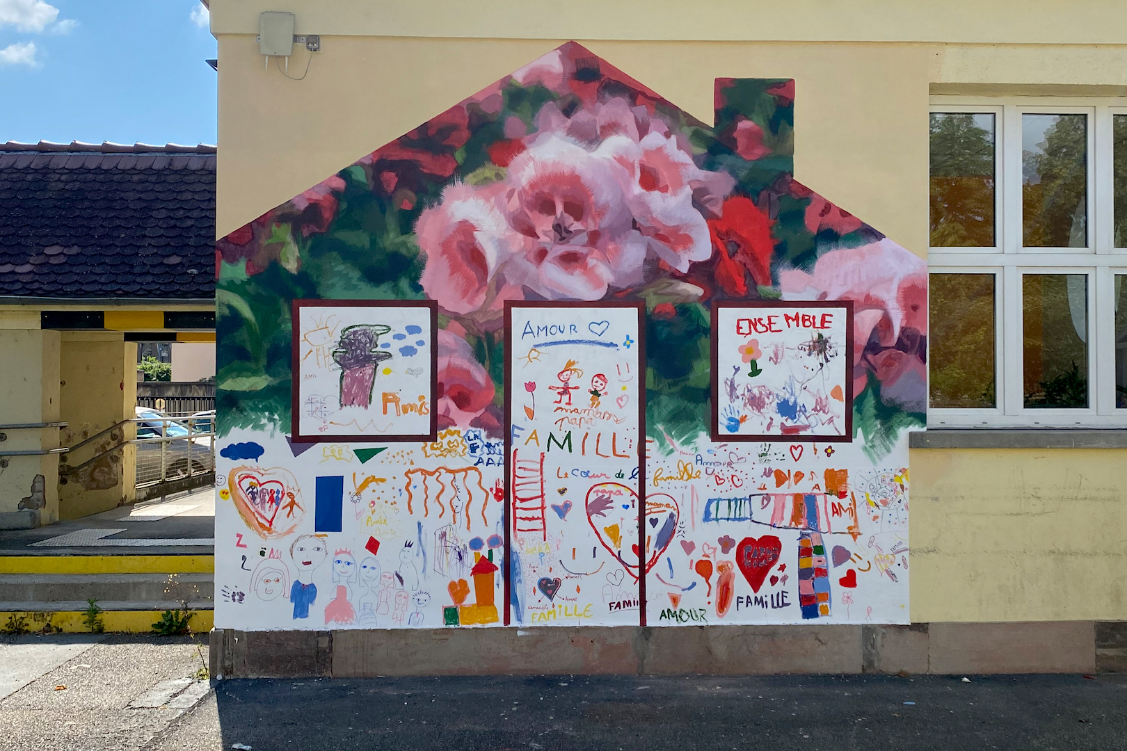 Fresque participative qui représente une forme de maison avec dedans des dessins d'enfants et une fleur de type geranium.